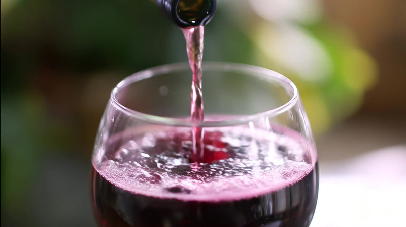 wine glass with red wine and a pouring spout set against a blurred background of greenery - Stock Video