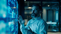 a man standing in an office environment gesturing with his hands while looking at a large screen various digital images and data - Stock Video