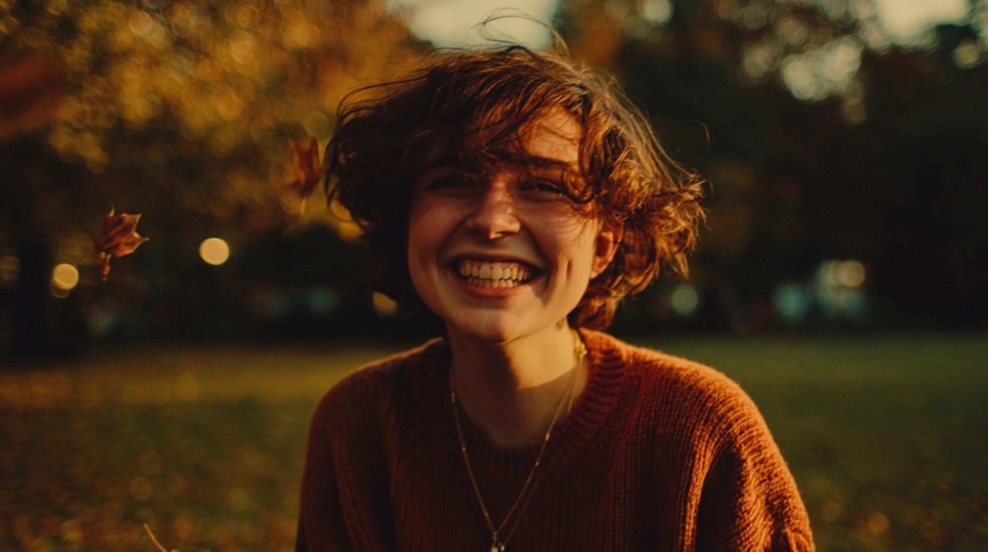a young woman is seated outdoors smiling and looking upwards as autumn leaves fall around her - Stock Video