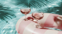 two champagne flutes on a pool float in a swimming pool with a tropical background palm trees and blue sky - Stock Video