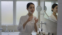 a woman in a bathrobe is standing at a bathroom sink applying makeup to her face - Stock Video