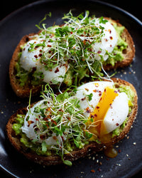 Avocado Toast With Fried Eggs Garnished With Microgreens And A Sprinkle Of Red Pepper Flakes Served On A Dark Plate — Stock Photo