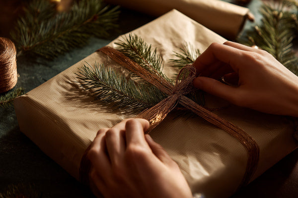 A Person Wrapping A Christmas Gift In Brown Paper With A Bow Surrounded By Pine Branches And Twinkling Lights — Stock Photo