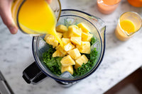 a person pouring orange juice into a blender filled with pineapple chunks and spinach leaves - Stock Video