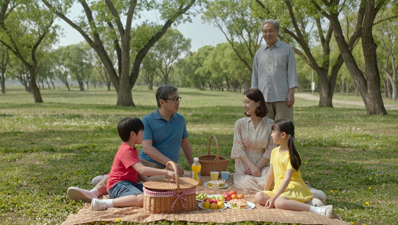 family of four sitting on blanket in park surrounded by fruit and drinks smiling towards camera - Stock Video