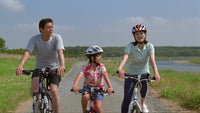 a family of three riding bicycles together in a rural setting with a clear sky and lush greenery in the background - Stock Video