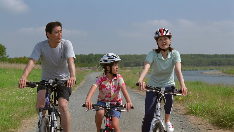 a family of three riding bicycles together in a rural setting with a clear sky and lush greenery in the background - Stock Video
