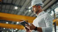 an industrial worker in a hard hat reviewing a tablet with both hands while standing in an indoor factory setting - Stock Video