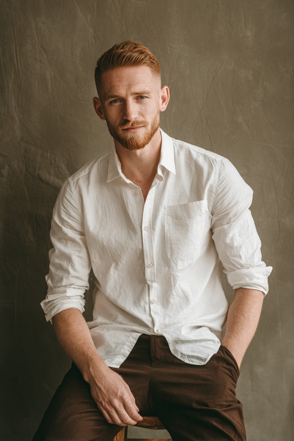 Man Posing For Portrait Wearing White Shirt And Brown Pants Seated On Stool Against Gray Background — Stock Photo