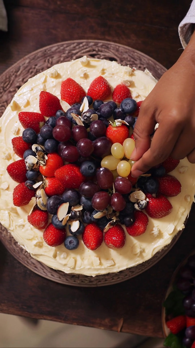 a person s hand reaching towards a cake topped with a variety of fruits including strawberries and grapes on a wooden table - Stock Video