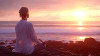 a serene sunset meditation scene with a woman sitting on rocks by the ocean silhouetted against the vibrant colors of the sky - Stock Video